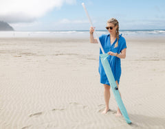 A woman in a blue dress setting up a Pop 'N Go Beach Umbrella on the sand at the beach for sun protection.