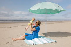 A woman in a blue dress setting up a Pop 'N Go Beach Umbrella on the sand at the beach for sun protection.