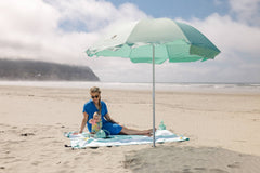 A woman in a blue dress setting up a Pop 'N Go Beach Umbrella on the sand at the beach for sun protection.