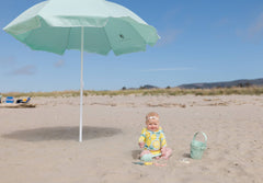 A woman in a blue dress setting up a Pop 'N Go Beach Umbrella on the sand at the beach for sun protection.