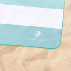The Coastline Beach Towel rolled up in aqua and white, showcasing its soft and absorbent fabric, perfect for summer.